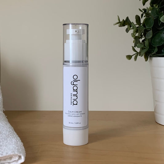 A white pump bottle of Olyanna Skincare Calm Cream stands on a light wooden surface against a beige wall, with a white towel on the left and a potted green plant on the right.