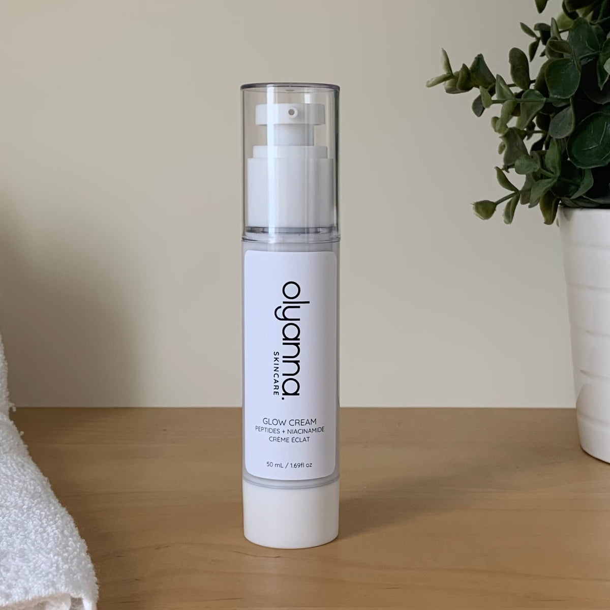 A bottle of Olyanna Glow Cream stands on a light wooden table against a beige wall, with a white towel on the left and a potted green plant on the right.