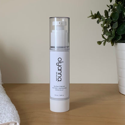 A bottle of Olyanna Glow Cream stands on a light wooden table against a beige wall, with a white towel on the left and a potted green plant on the right.
