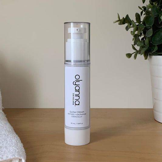 A bottle of Olyanna Glow Cream stands on a light wooden table against a beige wall, with a white towel on the left and a potted green plant on the right.