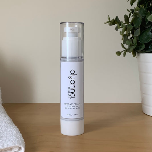 A bottle of Olyanna Skincare Hydrate Cream stands on a light wooden surface against a beige wall, with a white towel on the left and a potted green plant on the right.