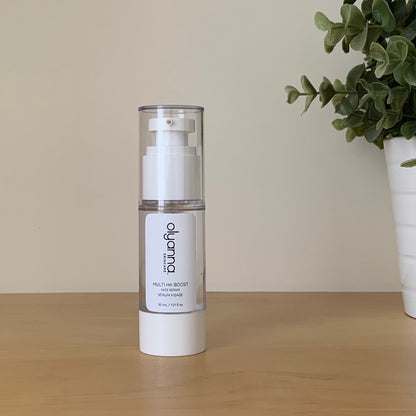 A white pump bottle of Olyanna Skincare Multi HA Serum stands on a light wooden surface against a beige wall, with a potted green plant on the right.