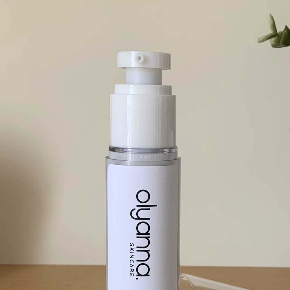 A white skincare pump bottle with a clear cap and black 'olyanna SKINCARE' branding, standing on a wooden surface against a beige wall.