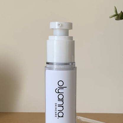 A white skincare pump bottle with a clear cap and black 'olyanna SKINCARE' branding, standing on a wooden surface against a beige wall.